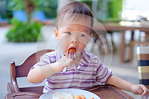 Asian 18 months / 1 year old toddler boy child sitting in high chair using fork eating whole cherry tomatoes