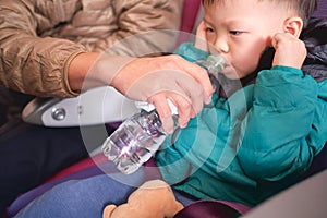 Asian 2 - 3 years old toddler boy child holding his aching ear and drinking water from bottle during flight on airplane. Little