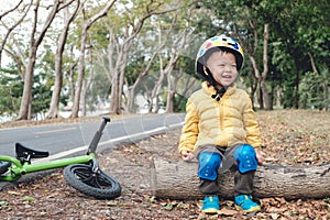 Asian 2 - 3 years old toddler boy child wearing safety helmet, sitting on a wooden log taking a break after riding his balance