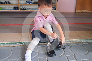 Asian 3 - 4 years old kindergarten kid in uniform sitting and concentrate on putting on children`s classics boys black leather