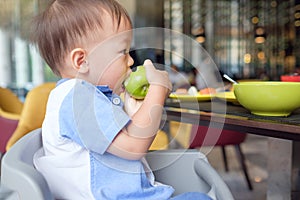 Asian 30 months / 2 years old toddler baby boy child sitting on high chair holding, biting, eating an unpeeled whole green apple a