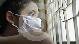 Asian adorable girl wearing protective mask looking out of window hopelessly during COVID-19 outbreak situation.