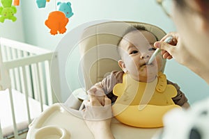 Asian Adorable little baby boy wearing yellow bib eating and sitting on high chair at home. Mother feeding pureed food to her son