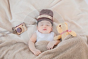 Asian adorable newborn baby wear brown knit hat deeply sleeping with beige blanket next to teddy bear and toy camera with safe and