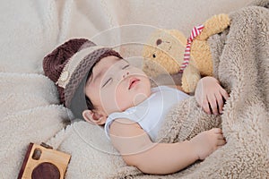 Asian adorable newborn baby wear brown knit hat deeply sleeping with beige blanket next to teddy bear and toy camera with safe and