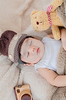 Asian adorable newborn baby wear brown knit hat deeply sleeping with beige blanket next to teddy bear and toy camera with safe and