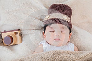 Asian adorable newborn baby wear brown knit hat deeply sleeping with beige blanket next to toy camera with safe and comfortable.