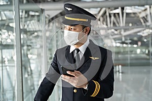 Asian Airliner pilot wearing face mask, standing in airport terminal. Young handsome captain using mobile phone chatting and