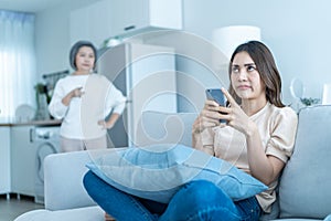 Asian angry senior mature woman shouting at stubborn fussy daughter. Young female sit on sofa use mobile phone chat, disobedient