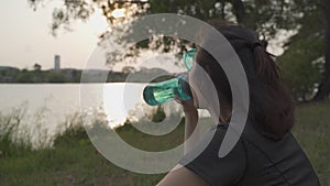 Asian athlete woman in green nature tree park and lake in evening sunset feeling thirsty drink water from bottle. energy drinks re