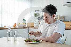 Asian attractive handsome male eating green salad in kitchen at home. Active young man sit on table, feel happy and enjoy
