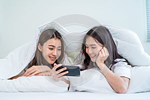 Asian attractive lesbian couple use mobile phone watch movie on bed. Beautiful sibling in pajamas lying down with blanket in