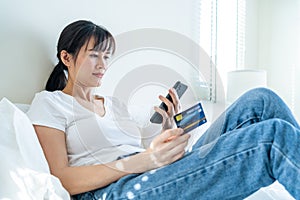 Asian attractive woman enjoy shopping online on bed at home in morning. Casual female lying in bedroom, holding credit card to