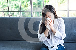 Asian attractive woman sitting on the sofa at home, is using tissue paper to wipe his nose because of coughing and sneezing due t