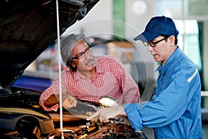 Asian automotive mechanic with blue uniform point to the car engine and explain about the problem to the customer in the garage