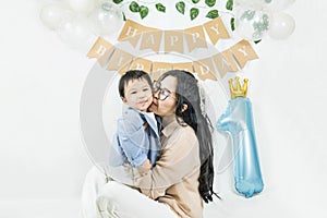 Asian baby boy celebrating first birthday,A mother hug and kiss her cheek son smiling on a floor with a minimal background,white