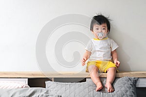 Asian baby with pacifier in her mouth is sitting on the bed in bedroom