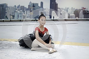 Asian ballerina dancer girl sitting on floor on rooftop with skyscraper city view, wearing her shoes, preparng for practice for