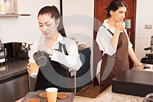 Asian barista women fill milk into takeaway hot coffee cup for customer while caucasian barista woman drinking hot black coffee in