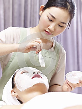 Asian beautician applying facial mask on face of young woman