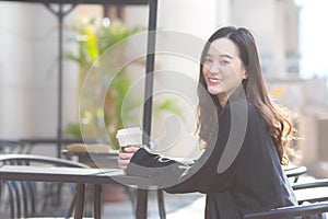 Asian beautiful femeal in a dark blue suit is sitting in a chair holding a coffee cup smiling looking at In front of the coffee