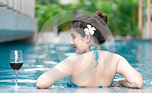 Asian beautiful girl smiling & drinking red wine  with white flower at the ear by wearing light blue swimming suit in the  pool. P