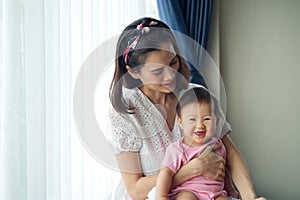 Asian beautiful mother holding her cute baby in her arms sitting near the window at home.