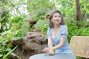 Asian beautiful woman in a blue dress is sitting happily smiling on the green garden and waterfall background