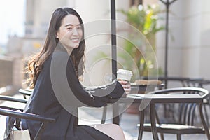Asian beautiful woman in a dark blue suit is sitting in a chair holding a coffee cup smiling looking at In front of the coffee