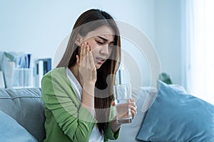 Asian beautiful woman feel terrible toothache after drink cold water. Attractive female sit on sofa in living room touching cheek