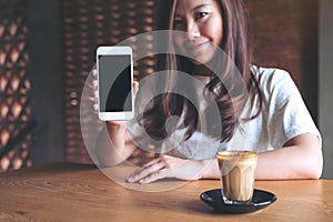 An asian beautiful woman holding and showing white mobile phone with blank black desktop screen with coffee cup on wooden table