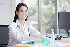 Asian beautiful young smiling female doctor wears rubber glove sitting in office at hospital. On table has a paper,face mask ,