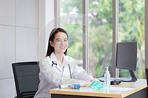 Asian beautiful young smiling female doctor wears rubber glove sitting in office at hospital. On table has a paper,face mask ,