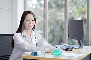 Asian beautiful young smiling female doctor wears rubber glove sitting in office at hospital. On table has a paper,face mask ,