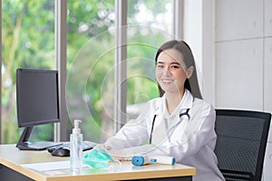 Asian beautiful young smiling woman doctor wears rubber glove sitting in office at hospital. On table has a paper,face mask ,