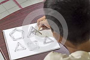 Asian boy learning and practicing to draw 3D shapes on drawing notebook on brown desk at home