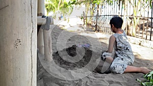 Asian boy playing alone enthusiastically digging sand with bare hands on the sandy shore. Bamboo pole obscures the view.