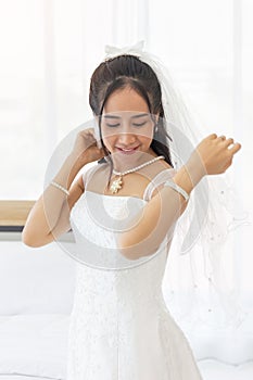 An Asian bride dressed in a white wedding dress stands in a cute and bright smile beside the bed in the room