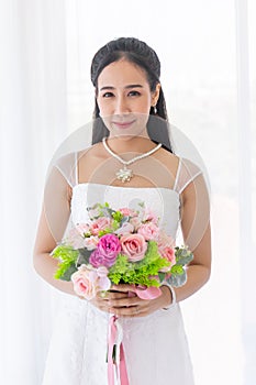 An Asian bride dressed in a white wedding dress stands smiling brightly in a hand holding a beautiful flower bouquet