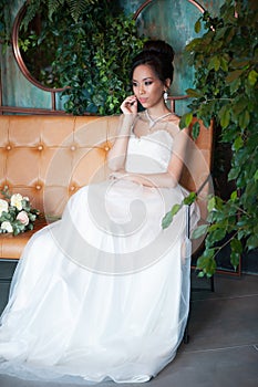 Asian bride with flowers in white dress sitting on sofa