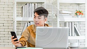 Asian business man sitting at desk with coffee cup and checking messages by mobile smartphone with laptop computer and calculator