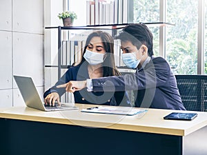 Asian business man and woman wearing suit and protective face masks using computer on desk, meeting and working together in office