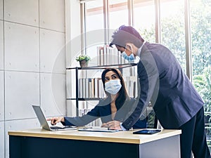 Asian business man and woman wearing suit and protective face masks using computer on desk, meeting and working together in office