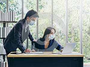 Asian business man and woman wearing suit and protective face masks using computer on desk, meeting and working together in office