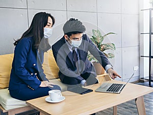 Asian business man and woman wearing suit and protective face masks using computer on desk, meeting and working together in office