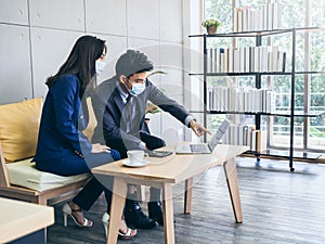 Asian business man and woman wearing suit and protective face masks using computer on desk, meeting and working together in office