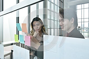 Asian business man and woman looking at paper note stick on glass board to discuss and working in office