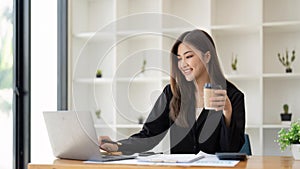 Asian business woman holding a cup of coffee at the desk Financial graph with desktop and laptop calculator for office