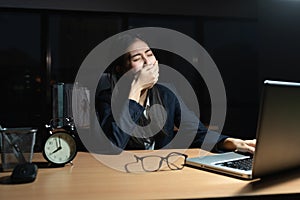 Asian business woman is sitting yawning with a hand covered her mouth, Laptop, on the desk, While the young woman works overtime