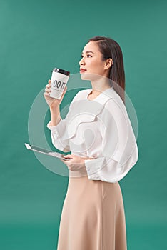 Asian business woman standing over  turquoise background, holding takeaway coffee, carrying laptop